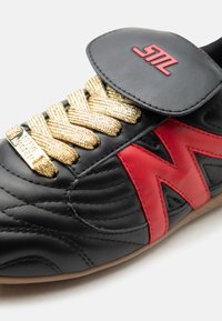 Μαύρο δερμάτινο αθλητικό παπούτσι με κόκκινο πλαϊνό γράμμα «Μ», κορδόνια με χρυσή γκλίτερ και μαύρο δερμάτινο λουράκι με κόκκινο λογότυπο «SM» σε λευκό φόντο.