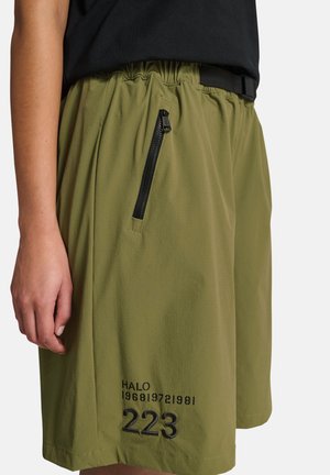 Persoon draagt olijfgroene korte broek met zwarte ritszak en zwarte bedrukte tekst "HALO 223" op de linker dij, gecombineerd met een zwart shirt.