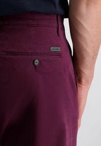 Bordeaux kleurige broek met een achterzak met knoopsluiting en een klein metalen logo label. Glad materiaal met een getailleerde pasvorm.