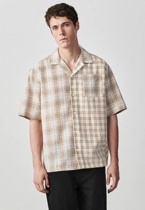 Junger Mann trägt ein beige-weiß kariertes kurzärmliges Hemd mit Brusttasche, steht vor einem schlichten hellen Hintergrund.