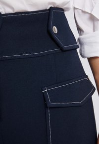 Granatowe spodnie z wysokim stanem, szerokim pasem, kieszeniami bocznymi, kontrastowym białym szwem oraz zapięciem na zatrzask w pasie.