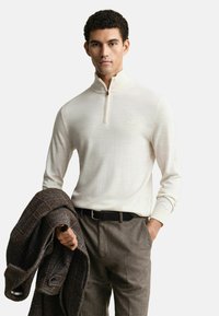 Junger Mann in einem cremefarbenen Pullover mit Viertelreißverschluss, braunen Wollhosen, schwarzem Gürtel, der einen dunkelbraunen karierten Mantel über einem Arm hält.