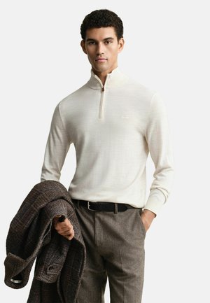 Jeune homme portant un pull crème à fermeture éclair quart, un pantalon en laine marron, une ceinture noire, tenant un manteau à carreaux brun foncé sous un bras.