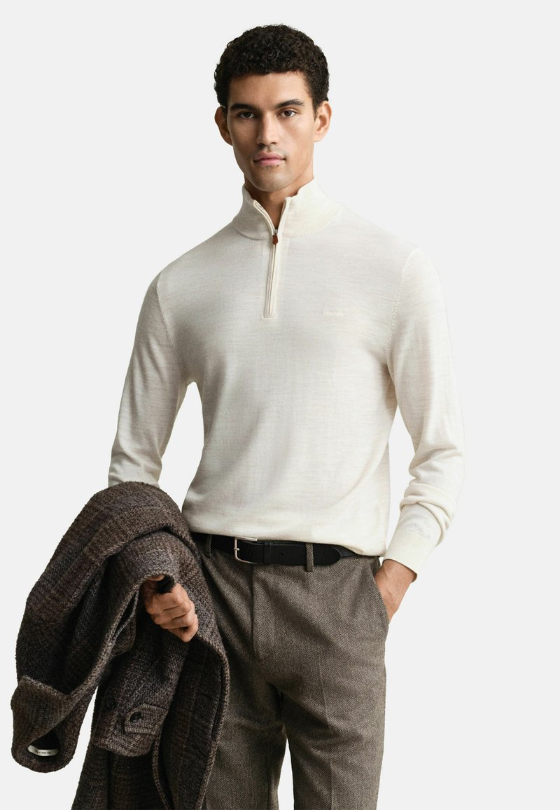 Junger Mann in einem cremefarbenen Pullover mit Viertelreißverschluss, braunen Wollhosen, schwarzem Gürtel, der einen dunkelbraunen karierten Mantel über einem Arm hält.