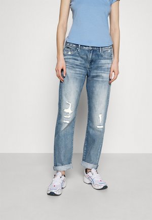 Personne portant un jean bleu clair retroussé et déchiré, des baskets blanches et un haut ajusté bleu clair à manches courtes, debout devant un fond uni.