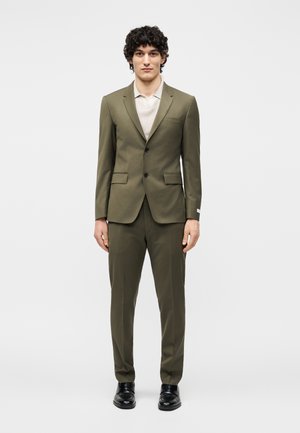 Νέος άνδρας που στέκεται φορεμένος με πράσινο-ελαιοχρώματο κοστούμι με δύο κουμπιά και τσέπες με καπάκια, άσπρο πουκάμισο με γιακά και μαύρα επίσημα παπούτσια σε απλό λευκό φόντο.