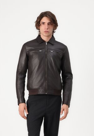 Man draagt een donkerbruine leren jas met ritszakken op de borst en een zwarte broek, staand tegen een effen witte achtergrond.