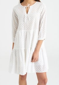 Vestido blanco de algodón perforado, de longitud hasta la rodilla, con mangas de tres cuartos, un escote redondo y un detalle de lazo en la parte frontal.