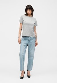 Jeune personne aux cheveux foncés portant un t-shirt gris à manches courtes, un jean bleu clair droit, et des talons hauts noirs, debout sur fond blanc.