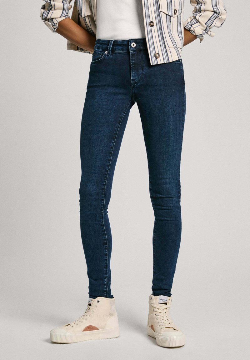Jeans skinny de color azul oscuro hechos de denim, con un diseño clásico de cinco bolsillos y una textura suave. Combinados con zapatillas altas blancas.