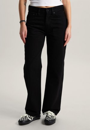 Kvinde iført sorte jeans med lige ben, hvid croptop og sort-hvide ternede snøresko, stående mod en ensfarvet baggrund.