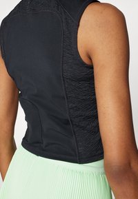 Gros plan sur le haut du dos et le bras d'une personne portant un haut sans manches noir texturé et une jupe plissée vert clair.