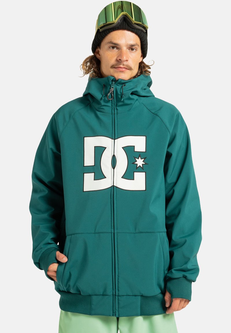 Man in een teal hoodie met groot wit DC-logo, zwarte beanie, skibril op het hoofd en lichtgroene broek, staande tegen een witte achtergrond.