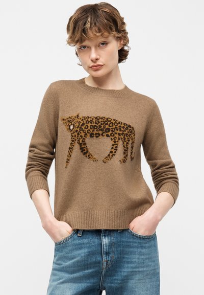 Jeune personne aux cheveux courts et bouclés portant un pull marron avec un motif de léopard en marche, associée à un jean bleu, les mains dans les poches.