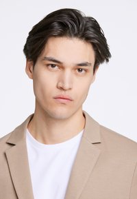 Beżowa marynarka na białym t-shircie; gładki materiał z klapami w kształcie litery V; model ma krótkie ciemne włosy i neutralny wyraz twarzy; jednolite białe tło.