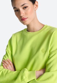 Zöld pulóver környakkal, bordázott mandzsettával és egy kis hímzett logóval a mellkason. Lágy, texturált anyagból készült.