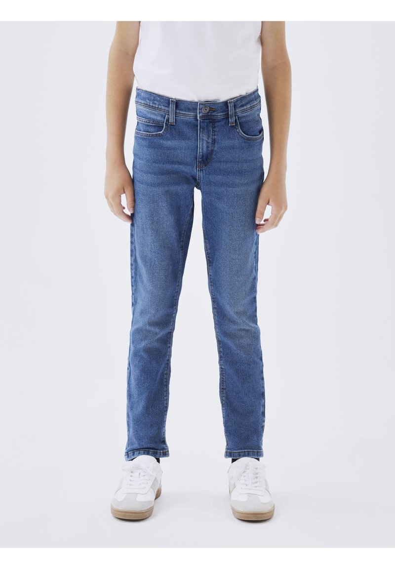 Enfant portant un jean slim bleu avec des poches avant et des baskets blanches, debout devant un fond clair uni.
