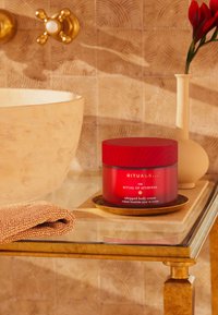 Punainen lasipurkki, jossa on puukansi ja etiketti "Rituals The Ritual of Ayurveda Whipped Body Cream". Vaaleanbeige pesulappu kultaisella tarjottimella sen vieressä.