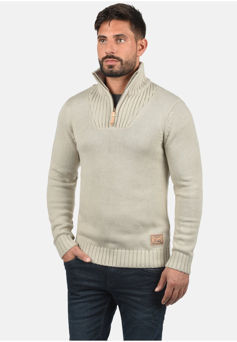 Beige gebreide trui met een hoge ribbelkraag, half-rits sluiting en een vierkante leren patch aan de zoom. Draag een donkere spijkerbroek.