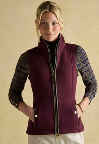 Maroone Fleece-Weste mit hohem Kragen und Reißverschluss. Sie verfügt über seitliche Taschen, goldfarbene Hardware-Details und ist über einem gemusterten blauen Oberteil getragen.