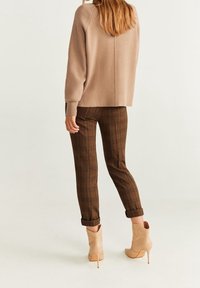 Femme portant un pull en maille beige, un pantalon ajusté à carreaux marron retroussé aux chevilles et des bottines à talons beiges, debout devant un fond uni.