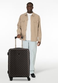 Zwarte koffer met een patroon op het oppervlak, stevige wielen en een uittrekbare handgreep, weergegeven naast een persoon in een beige shirt en lichte jeans.