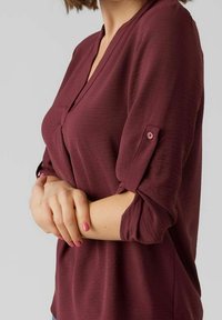 Blouse bordeaux avec un décolleté en V et des manches retroussées. Texture douce, avec un détail de bouton sur la manche. Tissu léger.