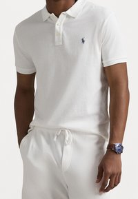 Polo en coton blanc avec col classique, patte de boutonnage à deux boutons et petit logo bleu. Porté avec un pantalon blanc et une montre-bracelet marron.