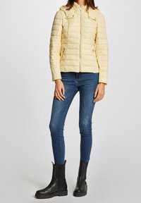 Padded yellow jacket with horizontal quilting, zip closure, and removable hood. Paired with blue jeans and black ankle boots.