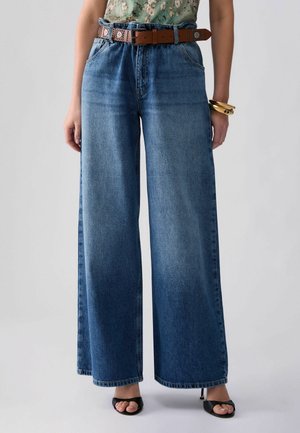 Kvinde iført højtaljede, vidde blå jeans, brun bælte, blomstret top, sorte åbne hæle og guldbaner på det ene håndled.
