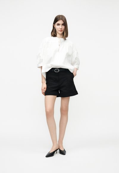 Jeune femme debout portant un chemisier blanc ample, un short noir ceinturé et des chaussures à petit talon noires, sur un fond blanc uni.
