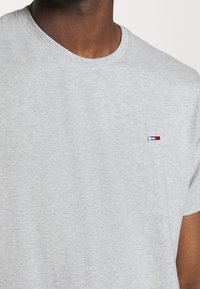 Šedé tričko s krátkým rukávem vyrobené z měkké tkaniny, s jemně vyšitým logem v červené, bílé a modré barvě na levé části hrudi.