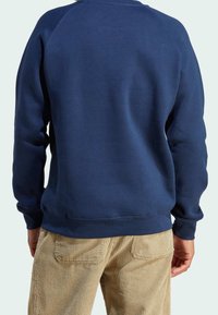 Person klädd i en enkel marinblå sweatshirt och beige corduroybyxor, visad bakifrån mot en ljus bakgrund.