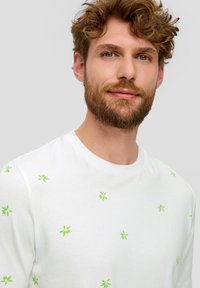 Biała bawełniana koszulka z małymi zielonymi wzorami liści rozmieszczonymi na tkaninie, okrągły dekolt i krótkie rękawy.