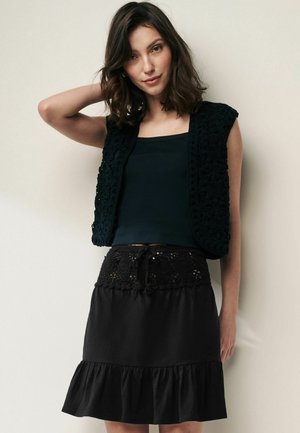 Gilet en crochet noir sur un débardeur sombre, associé à une jupe noire avec une taille élastique en dentelle et un bas à volants.