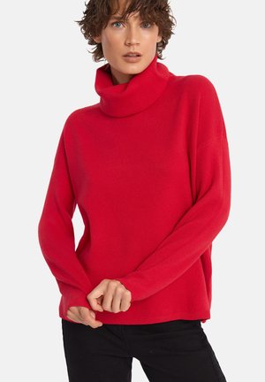 Czerwony sweter z kołnierzem typu golf o długich rękawach, o miękkiej teksturze, luźnym kroju i prążkowanych mankietach. Nosi się go na czarnych spodniach.