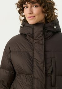 Brown puffer jacket with a high collar, featuring a zippered pocket and drawstring details. Made of smooth, quilted material.