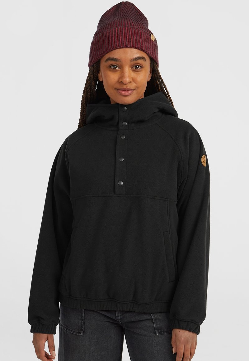 Woman wearing maroon knit beanie, black hooded pullover with snap buttons, and dark jeans, standing against plain light background.