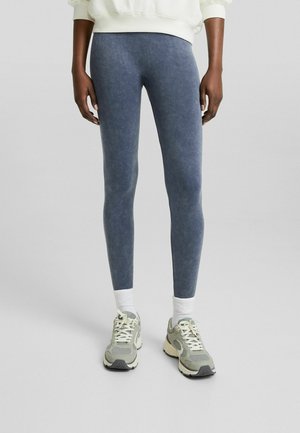 Strakke, donkerblauwe leggings met een gladde textuur, gecombineerd met lichtgrijze sneakers en witte enkelsokken. Eenvoudig en figuuraccentuerend ontwerp.