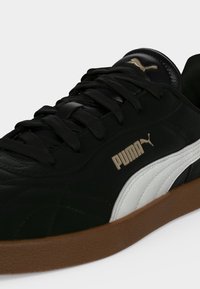 Μαύρο sneaker Puma με λευκή πλαϊνή λωρίδα, καφέ σόλα, μαύρα κορδόνια και χρυσό λογότυπο Puma στο πλάι και στη γλώσσα.