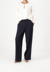 Vrouw die een crèmekleurige blouse met lange mouwen en een keyhole-halslijn draagt, samen met zwarte wijde broekspijpen, staat met handen in de zakken.