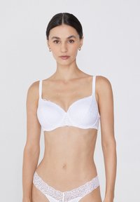 Witte kanten bh met gepolsterde cups, verstelbare bandjes en gekartelde randdetails. Gladde textuur met een bloemenpatroon over de stof.
