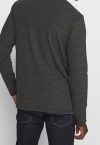 Graues Langarmshirt mit Rundhalsausschnitt, aus einem strukturierten Stoff gefertigt. Das Shirt wird mit dunklen Jeans kombiniert, die minimale Details aufweisen.