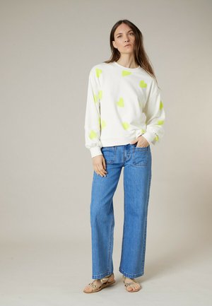 Femme debout portant un sweat-shirt blanc avec des cœurs jaune néon, un jean bleu avec des poches avant, et des sandales beiges sur un fond uni.