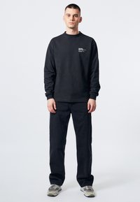 Sudadera negra de manga larga con cuello redondo, combinada con pantalones negros. La sudadera presenta texto y logotipos en el pecho.
