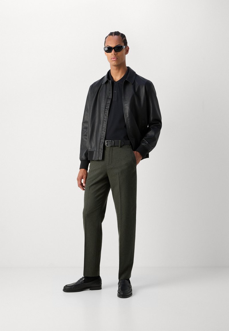 Schwarze Lederjacke über einem dunklen Polo-Shirt, kombiniert mit olivgrünen Hosen und schwarzen Slippern. Die Accessoires umfassen schwarze Sonnenbrillen.