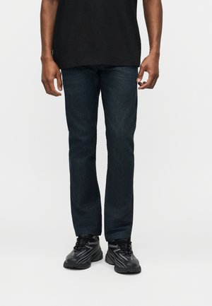 Uomo che indossa jeans dritti blu scuro, sneaker nere con dettagli grigi e una maglietta nera, in piedi contro uno sfondo bianco.