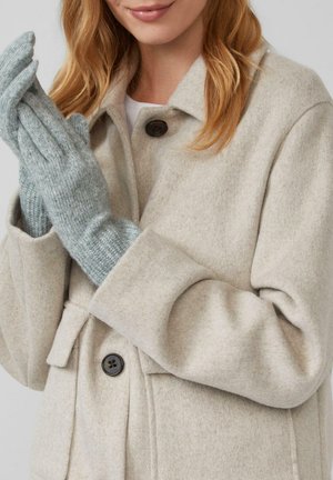 Vrouw in een beige wollen jas met zwarte knopen, die lichtgrijze gebreide handschoenen aantrekt, met schouderlang golvend blond haar.