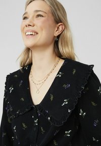 Chemise en velours côtelé noir ornée de broderies florales blanches et violettes, avec un col à volants et trois colliers en or superposés avec pendentifs.