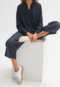 Personne portant un chemisier bleu marine, un pantalon gris raccourci et des baskets blanches assise en tailleur sur un cube blanc sur fond blanc.